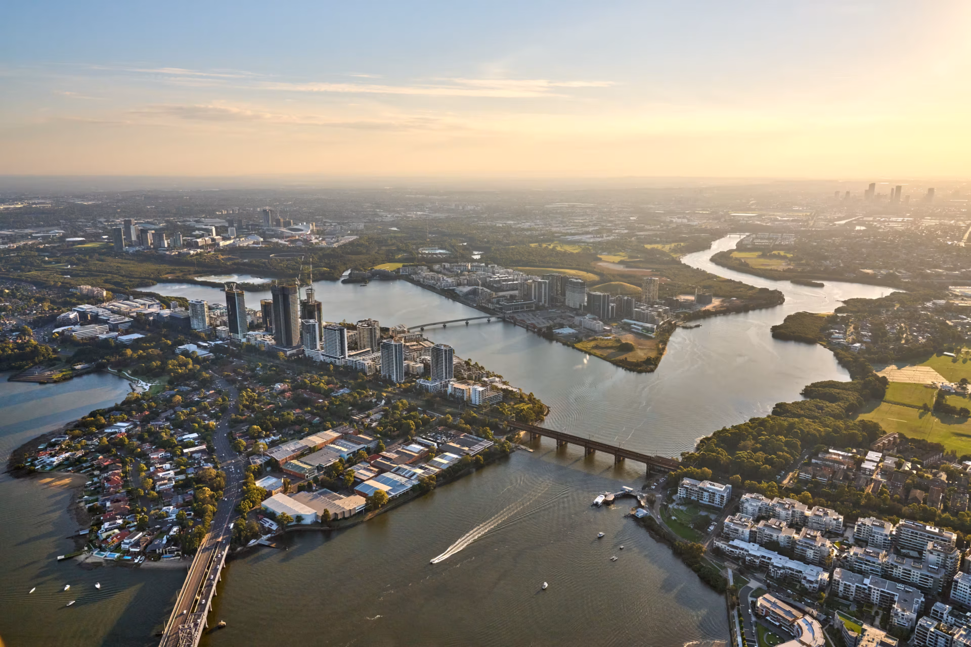 Helicopter aerial view of Rhodes and the Parramatta River at sunrise, showing the John Whitton Bridge, high-rise apartments, and a boat leaving a wake on the water under a warm golden glow.