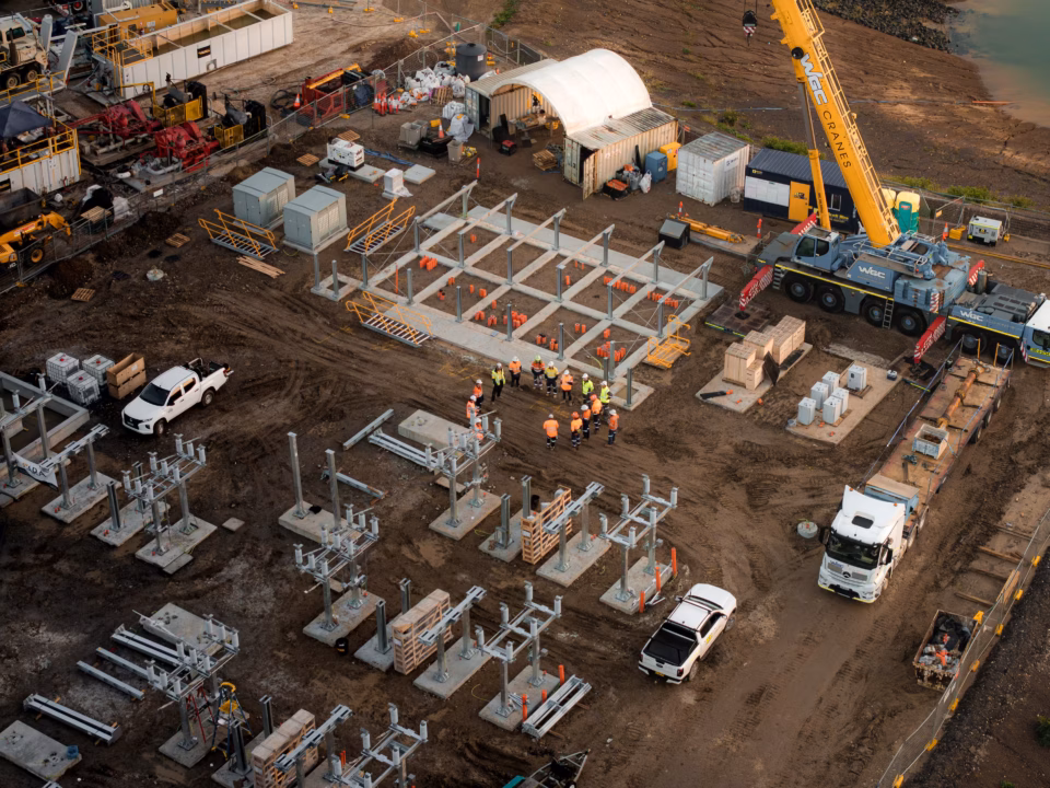 An aerial view of a construction site for an electrical substation, featuring a group of workers in high-visibility vests gathered for a meeting amidst steel frameworks, heavy machinery including a yellow crane, and various construction equipment on a dirt ground.